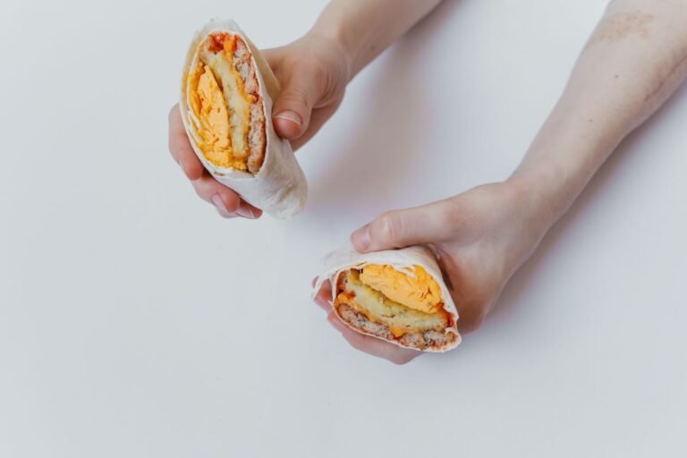 Classic Sausage, Egg, and Cheese Breakfast Burritos — A stack of foil-wrapped breakfast burritos next to one cut open showing sausage, egg, and melted cheese